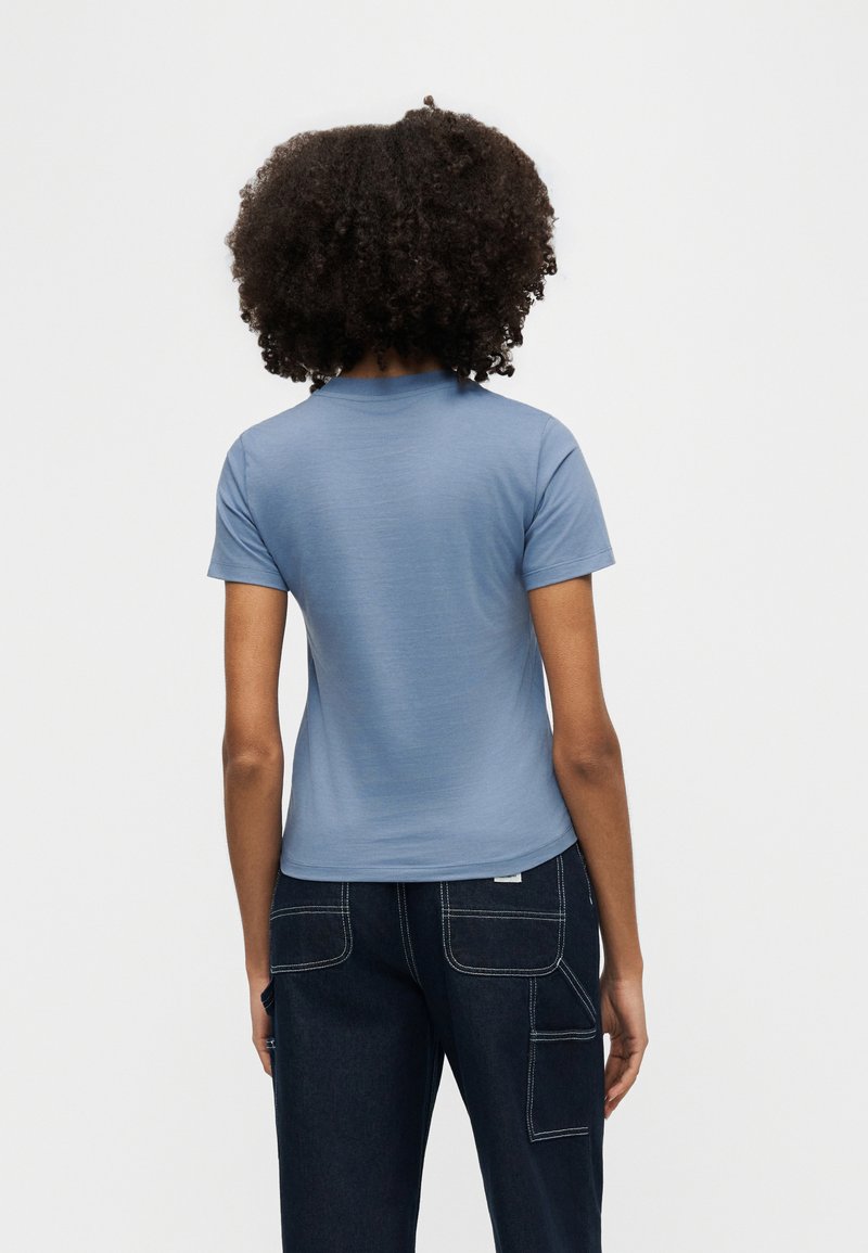 Eine Person mit lockigem Haar trägt ein hellblaues Kurzarmhemd und dunkelblaue Jeanshosen mit mehreren Taschen, schaut von hinten vor weißem Hintergrund.