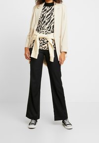 Blazer beige sopra un top a fantasia nera e crema, stretto in vita con una cintura crema. Pantaloni flared neri e sneaker nere completano il look.