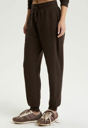 Osoba w ciemnobrązowych spodniach dresowych z sznurkiem w talii oraz brązowych, sznurowanych trampkach z beżowymi akcentami, stojąca na jasnym tle.