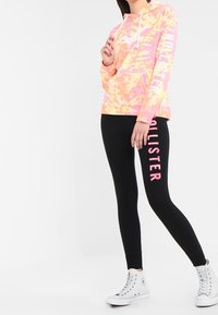 Osoba ubrana w różowo-żółtą bluzę tie-dye i czarne legginsy z napisem "Hollister" w różowych literach, z białymi trampkami za kostkę.