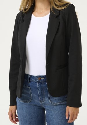 Frau trägt einen schwarzen Blazer über einem weißen Oberteil, kombiniert mit blauen Jeans, gezeigt von den Schultern bis zur Hüfte vor einem einfarbigen Hintergrund.