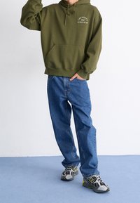 Oliwkowa bluza z kapturem z przednią kieszenią i sznurkiem do ściągania, w połączeniu z jasnoniebieskimi prostymi dżinsami i szarymi trampkami New Balance.