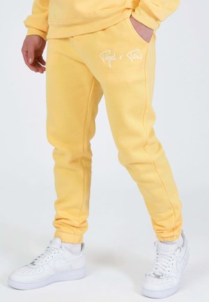 Personne portant un pantalon de jogging jaune avec le texte « Project x Paris » sur la poche et des baskets blanches, mains dans les poches, debout devant un fond uni.