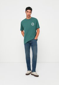 Tânărul stă cu mâinile în buzunare, purtând un tricou grafic verde, blugi albaștri și adidași albi, pe un fundal alb simplu.