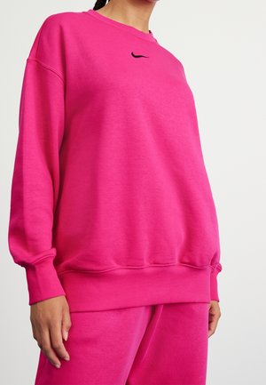 Kobieta ubrana w jasnoróżową, za dużą bluzę i pasujące spodnie z małym czarnym logo Nike na piersi.