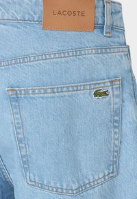 Shorts in denim chiaro con etichetta in pelle marrone che presenta la scritta "LACOSTE" e un piccolo logo a forma di coccodrillo verde sulla tasca posteriore.