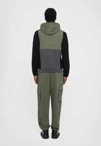Sweatshirt com capuz em verde oliva, cinza e preto; calças cargo com detalhes em verde nas laterais; material em algodão; ajuste relaxado; detalhes de costura visíveis.