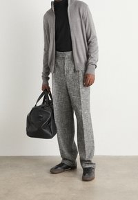 Cardigan grigio con zip sopra una camicia nera, abbinato a pantaloni grigi a gamba larga e dalla trama particolare. Borsa nera e scarpe scure completano l'outfit.