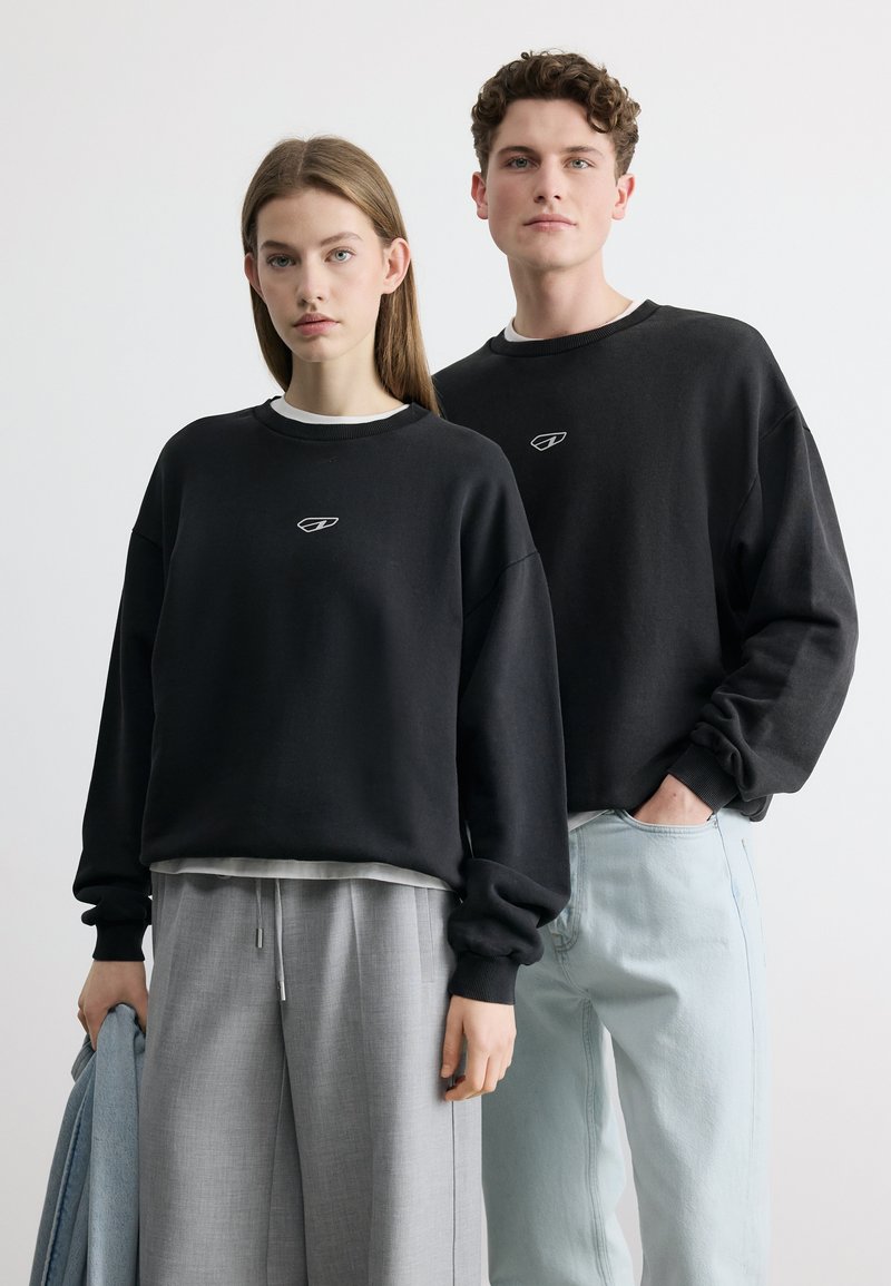 Νεαρή γυναίκα και άνδρας φορούν μαύρα φούτερ με λογότυπο, στέκονται μπροστά σε λευκό φόντο, η γυναίκα κρατά ένα τζιν μπουφάν.