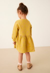 Vestido amarillo con patrones de arcoíris, mangas largas y falda acampanada. La niña lleva botas beige con acentos de volantes rosas.