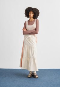 Vaalean värinen kerroksittainen maksimihame, jossa on kiristysnyöri vyötäröllä ja oranssiraidallinen yksityiskohta. Yhdistetty istuvaan pitkähihainen toppiin ja tennareihin.