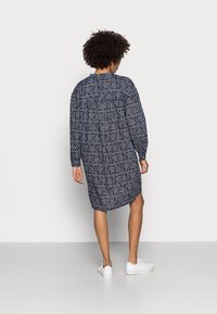 Robe bleu marine à manches longues avec un motif paisley, coupe décontractée, détail froncé au dos et baskets blanches. Texture de tissu lisse.