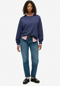 Granatowy, duży sweter zakładany na jasnoróżową, w paski koszulę, w połączeniu z niebieskimi dżinsami i czarnymi loafersami. Miękka tekstura, luźny krój.