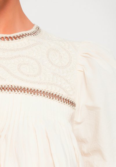 Close-up of white blouse with puffed sleeve, intricate embroidery, chain detail at neckline and yoke, and fine pleats below yoke.