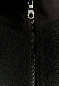 Schwarze Fleecejacke mit Reißverschluss und strukturierter Oberfläche, ausgestattet mit einem robusten Metallreißverschluss und hohem Kragen. Oben glatte Stoffakzente.
