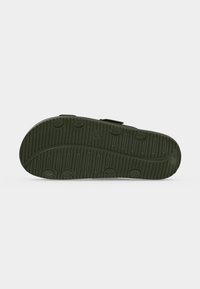 Semelle de sandale vert foncé avec des grips circulaires et des motifs de bande de roulement verticale sur fond blanc uni.