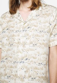 Camicia a maniche corte, con bottoni, di colore crema con un motivo floreale beige e blu. Presenta un colletto a scollo a V e una superficie liscia.