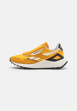 Zapatilla Reebok amarilla y blanca con acentos negros, diseño con cordones y suela texturizada mostrada de perfil sobre un fondo blanco.