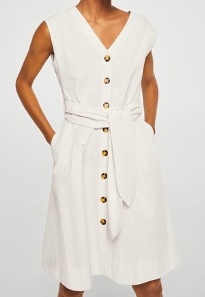 Ærmeløs hvid kjole med V-hals, forreste knapper i skildpaddeskaller, sidelommer og et bredt stofbælte bundet i taljen.