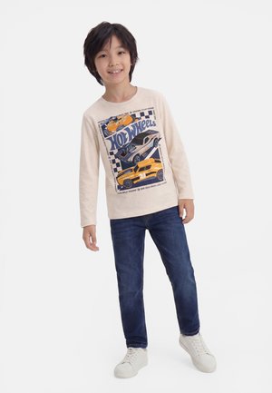 Garçon souriant portant un t-shirt crème à manches longues Hot Wheels, un jean bleu et des baskets blanches, debout devant un fond blanc.