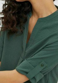 Chemise verte à manches courtes avec un décolleté en V profond, poignet de manche boutonné et un tissu doux et texturé. Couleur riche et coupe décontractée.