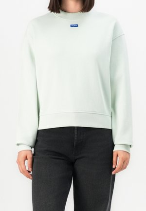 Vrouw draagt een lichtgroene sweater met ronde hals en een klein blauw "HUGO" logo op de borst, gecombineerd met zwarte jeans met een hoge taille.