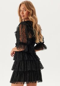 Svart spetsklänning med lager av volanger, genomskinliga puffärmar och rynkade detaljer vid halsringningen. Bakdelen har en nyckelhålsutskärning.