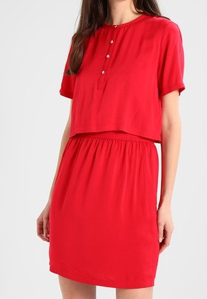 Woman wearing a short-sleeve red dress with a buttoned placket and a gathered elastic waistband, standing against a plain background.