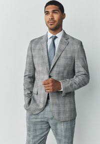 Traje de cuadros gris con chaqueta de un solo botón y pantalones a juego. Camisa azul claro y corbata de lunares añaden contraste. Ajuste liso y entallado.