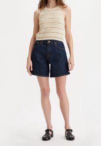 Beige knit halter top with open patterns and dark denim high-waisted shorts. Black strappy sandals complete the look.