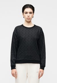 Sweatshirt preta com padrão texturizado de formas "A" entrelaçadas, punhos canelados e decote redondo. Usada com calças de cor clara.