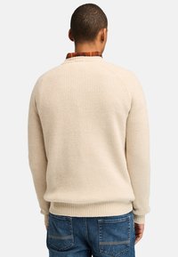 Maglione beige a coste con maniche lunghe, girocollo, trama strutturata e vestibilità comoda, indossato sopra una camicia a quadri.