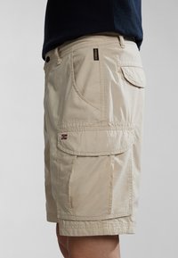Beige cargo šortky vyrobené z lehkého materiálu, s více kapsami, stehenním pasem a malým vyšitým logem.