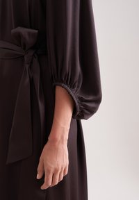 Dunkelbraunes Kleid mit lockerer Passform, elastischen Puffärmeln und einem gebundenen Taillengürtel. Glattes Material mit einer weichen Textur.