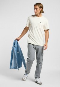 Camiseta corta de manga en crema con un bolsillo, pantalones cargo grises con bolsillos laterales y una chaqueta de mezclilla azul clara sostenida en la mano. Zapatillas blancas con rayas negras.