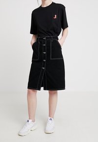 Falda de mezclilla negra con cierre delantero de botones y bolsillos, cintura alta, detalles de costura blanca, combinada con una camiseta negra y zapatillas blancas.