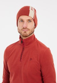 Berretto lavorato a maglia color ruggine con un accento crema. Pullover in pile caldo e morbido con mezza zip e dettaglio del logo sul colletto, completando il look.