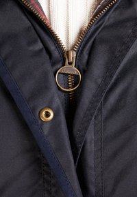 Navy waterdichte jas met gouden accenten, voorzien van een ritssluiting en een geruite voering. Zachte textuur en een knopelement aan de zijkant.