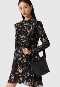 Vestido negro con estampado floral y volantes, con cuello alto y mangas largas. Combinado con un bolso tote de cuero negro.