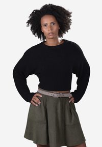 Woman with curly hair wearing a black knit sweater and olive green skirt with beige belt, standing with hands on hips, neutral expression.