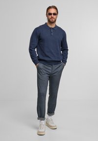 Mann in einem navyblauen langärmeligen Poloshirt, grauen hochgekrempelten Hosen, weißen Sneakers und Sonnenbrille, steht mit Händen in den Taschen vor einem grauen Hintergrund.