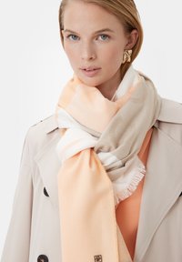Beżowo-miękki szalik w odcieniu pomarańczowym z delikatnym wzorem, z frędzlami, nałożony na jasną kurtkę, prezentowany na jednolitym tle.