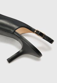 Svart skinne slingback-hæl med spiss tå, slankt design, gullfarget spenne og avsmalnende høy hæl.