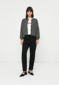 Donna in piedi contro un muro bianco, indossa un cardigan grigio con bottoni, una camicia bianca con stampa di labbra rosse, pantaloni neri e scarpe Mary Jane nere.