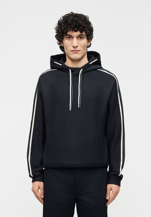 Jeune homme aux cheveux bouclés portant un sweat à capuche noir avec des bandes blanches et vertes sur les manches et un pantalon foncé assorti.