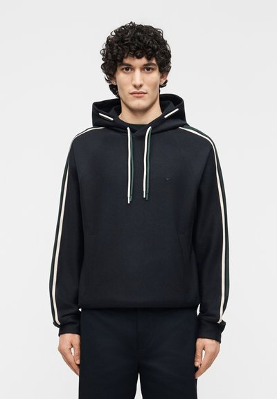Giovane uomo con capelli ricci che indossa una felpa nera con strisce bianche e verdi sulle maniche e pantaloni scuri coordinati.