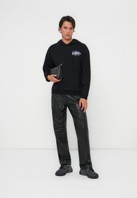 Haut à capuche noir avec une poche avant et un design graphique ; associé à un pantalon en cuir noir et des baskets noires ; tenant un petit sac noir.