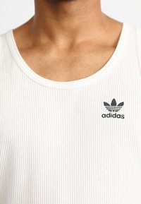 Camiseta sin mangas blanca de canalé con escote redondeado, que presenta un pequeño logo negro de Adidas en el lado izquierdo. Textura suave y diseño ajustado.