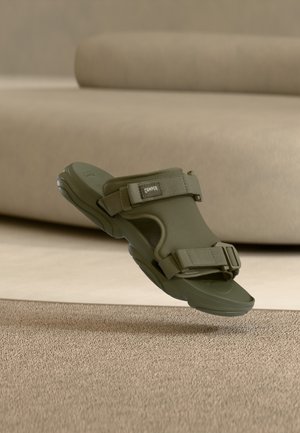 Olivengroene open-teen sandaal met verstelbare bandjes en gevoerde zool, zwevend boven een beige tapijt met textuur en een bijpassende bank op de achtergrond.