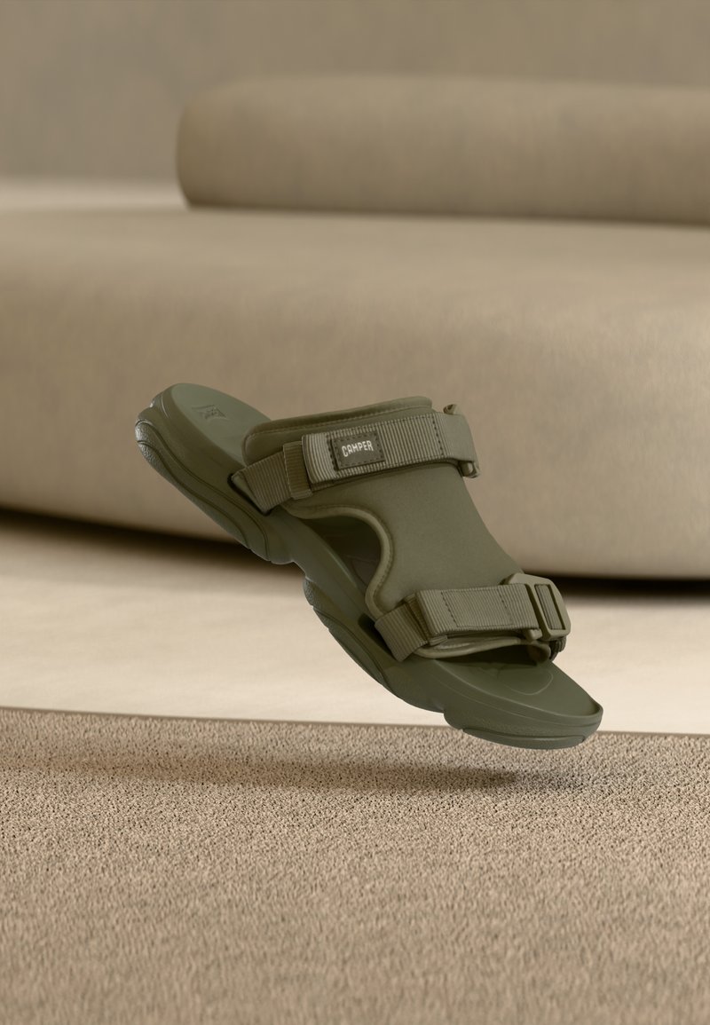 Sandale kaki à bouts ouverts avec brides ajustables et semelle rembourrée, flottant au-dessus d'un tapis beige texturé et d'un canapé assorti.